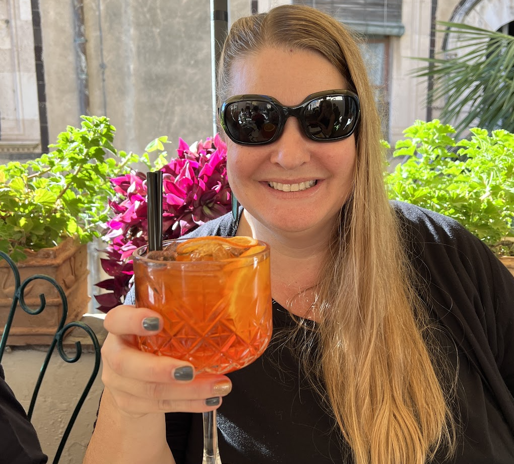 Lisa Sharples smiling outdoors and raising an orange cocktail